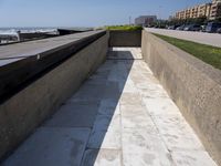 a cement bench on the side of a road next to the ocean waves and buildings