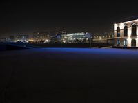 an empty basketball court next to a city at night time, at a distance is a neon lit skyscraper