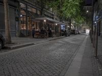 a city street at night with many parked cars on it and people sitting outside a small outdoor cafe in the background