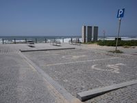 an empty parking lot near the water at the beach, and in the foreground is a blue street sign with numbers written in concrete and a circle
