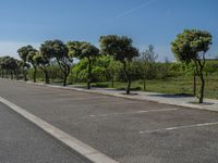 a road with trees in the parking lot of it next to a grass field and a fence