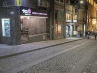 a street with a brick sidewalk at night time with cars parked outside the business and shops