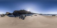 a sandy beach with rocks and water with blue skies in the background and sun streaming through