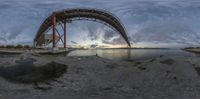 a view of the sunset under a large metal bridge over water and a sand beach