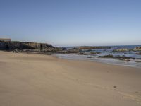 some people walking on the sand and sea water and rocks with a building in the background