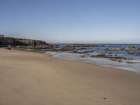 some people walking on the sand and sea water and rocks with a building in the background