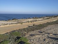 people walking along the road on the beach near the ocean waves and sand hills and scrubland