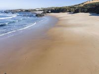 a sandy beach with waves washing on it and a steep hill in the distance next to water