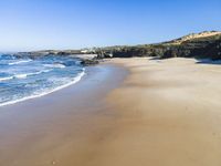 a sandy beach with waves washing on it and a steep hill in the distance next to water