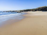 a sandy beach with waves washing on it and a steep hill in the distance next to water