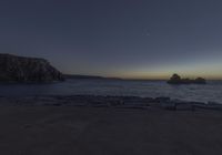 a clear blue ocean with some rocks in it near a cliff at dusk with the moon rising