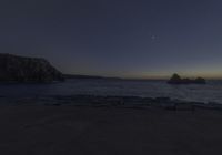 a clear blue ocean with some rocks in it near a cliff at dusk with the moon rising