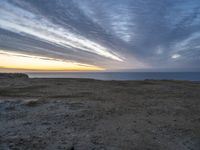 a lone bench is near the ocean during sunset, with an open sky behind it