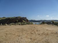 dirt field with wooden posts near rocks and mountains with a blue sky above it with clouds