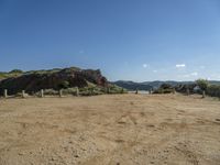 dirt field with wooden posts near rocks and mountains with a blue sky above it with clouds