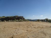 dirt field with wooden posts near rocks and mountains with a blue sky above it with clouds