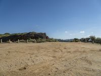 dirt field with wooden posts near rocks and mountains with a blue sky above it with clouds