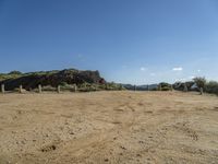 dirt field with wooden posts near rocks and mountains with a blue sky above it with clouds