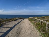 a wooden fence and rail on a dirt road beside the water with the sky in the background