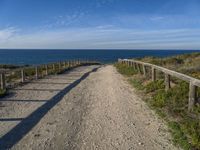 a wooden fence and rail on a dirt road beside the water with the sky in the background