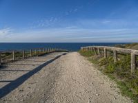 a wooden fence and rail on a dirt road beside the water with the sky in the background