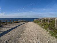 a wooden fence and rail on a dirt road beside the water with the sky in the background