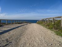 a wooden fence and rail on a dirt road beside the water with the sky in the background