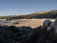 a sandy beach filled with lots of rocks near a shore covered with water, along side a hill and forest