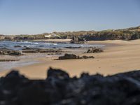 a sandy beach filled with lots of rocks near a shore covered with water, along side a hill and forest