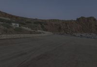 a truck drives along the side of the road near a cliff with houses, cars and mountain in the background