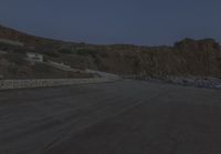 a truck drives along the side of the road near a cliff with houses, cars and mountain in the background