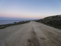 a dirt road near a body of water and hills at sunset with a lighthouse in the background