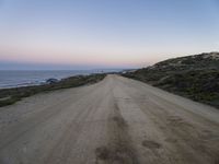 a dirt road near a body of water and hills at sunset with a lighthouse in the background