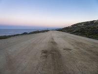 a dirt road near a body of water and hills at sunset with a lighthouse in the background