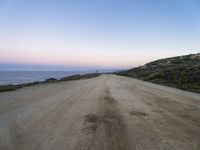 a dirt road near a body of water and hills at sunset with a lighthouse in the background