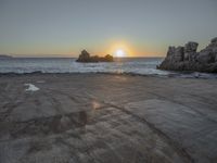 a sandy beach with a body of water near the sun setting in the distance, and a rock cliff in the middle of the ocean