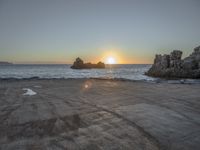 a sandy beach with a body of water near the sun setting in the distance, and a rock cliff in the middle of the ocean