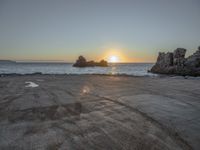 a sandy beach with a body of water near the sun setting in the distance, and a rock cliff in the middle of the ocean