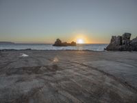 a sandy beach with a body of water near the sun setting in the distance, and a rock cliff in the middle of the ocean