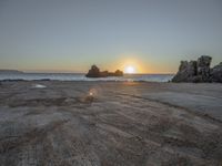 a sandy beach with a body of water near the sun setting in the distance, and a rock cliff in the middle of the ocean