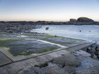 a fire hydrant next to the ocean with rocks and water surrounding it with boats on the water