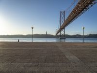 a large bridge is crossing the water at sunset time as seen from the sidewalk near the water