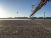 a large bridge is crossing the water at sunset time as seen from the sidewalk near the water