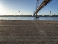 a large bridge is crossing the water at sunset time as seen from the sidewalk near the water