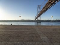 a large bridge is crossing the water at sunset time as seen from the sidewalk near the water