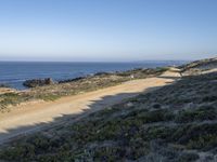 a long empty road and dirt path near the ocean on a clear day with a few people sitting on it