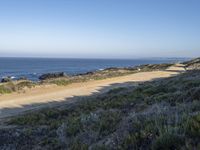 a long empty road and dirt path near the ocean on a clear day with a few people sitting on it