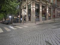 a cobble stone street at night, lined with parked cars and trees along the sidewalk