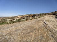 a dirt road with fence around it leading to the ocean, and mountains in the background