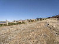 a dirt road with fence around it leading to the ocean, and mountains in the background